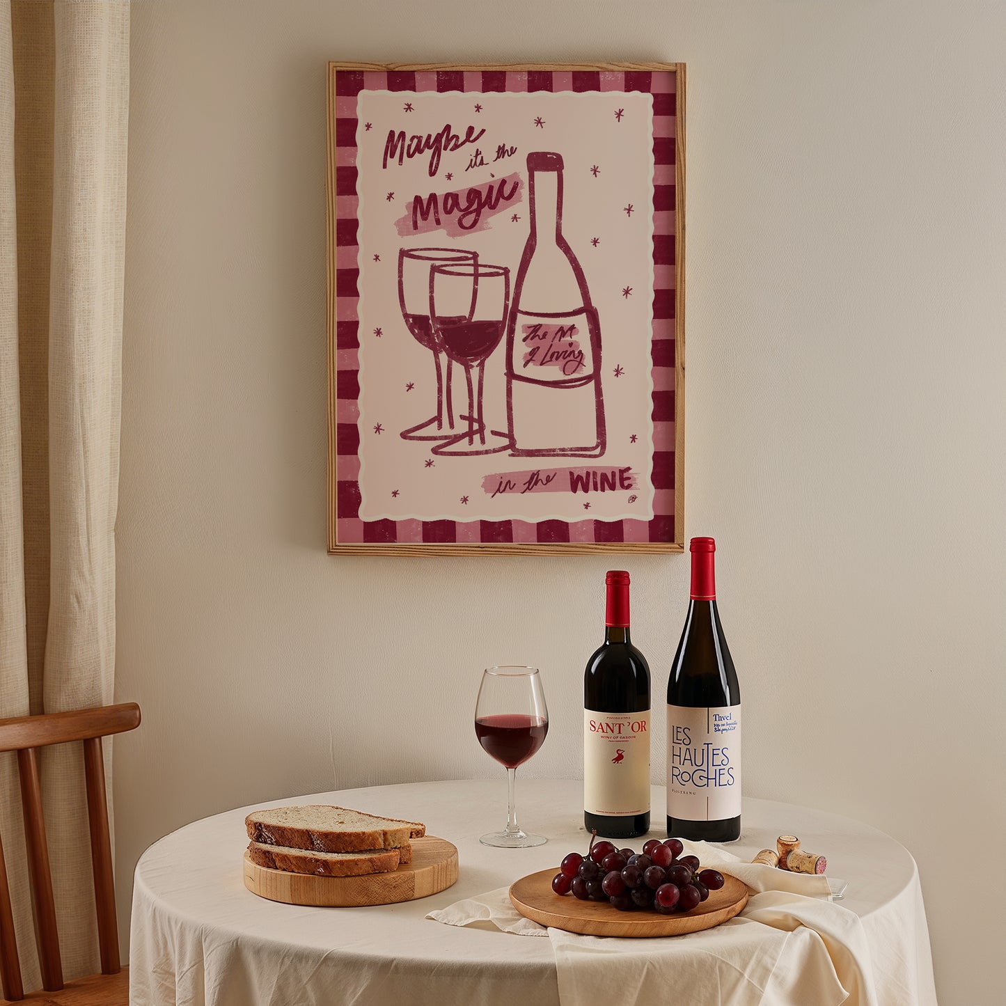framed artwork of a bottle of wine and two wine glasses with a pink checked border on the wall above a dining table with red wine and charcuterie