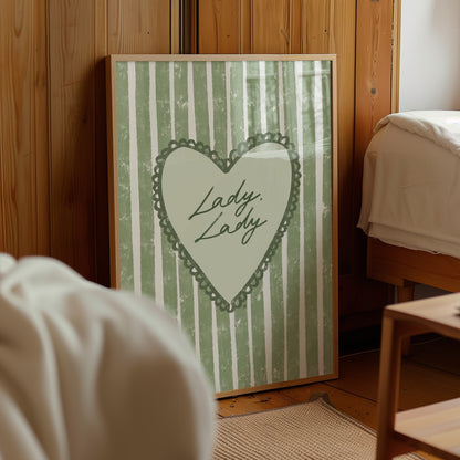 Framed artwork with a lacy sage green heart design and lady lady lyrics text on a wooden wall.